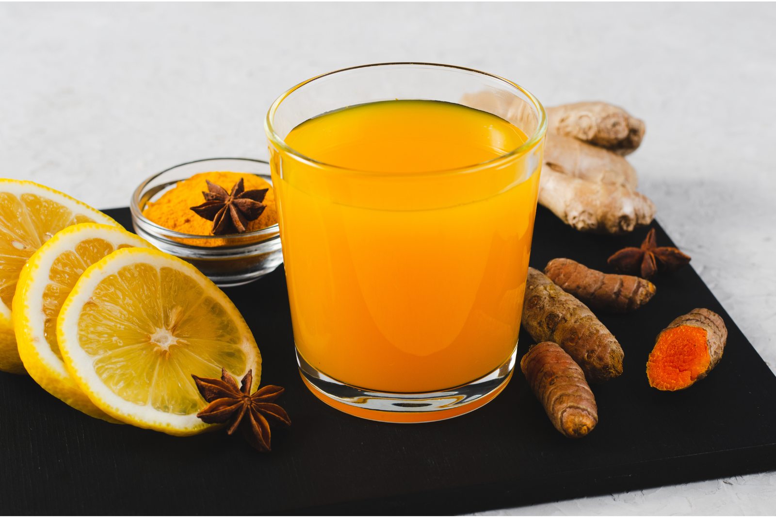 A shot glass of vibrant orange turmeric juice is centered on a black surface. Surrounding the shot glass are slices of fresh lemon, whole turmeric roots, ginger pieces, and anise stars. A small bowl with turmeric powder is also present, contributing to the colorful and aromatic display. An immunity superhero. Best to consume first thing in the morning or prior to bed. 