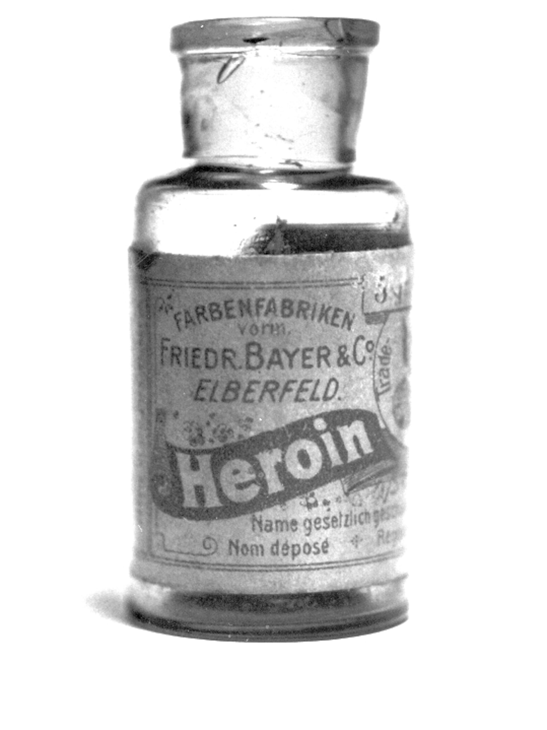 A vintage, cylindrical glass bottle with a cork stopper labeled "Heroin" from Farbenfabriken vorm. Friedr. Bayer & Co. Elberfeld, often used in early prescriptions. The label mentions trademarks in multiple languages and includes elaborate text detailing the product's name and origin. The image is in black and white.