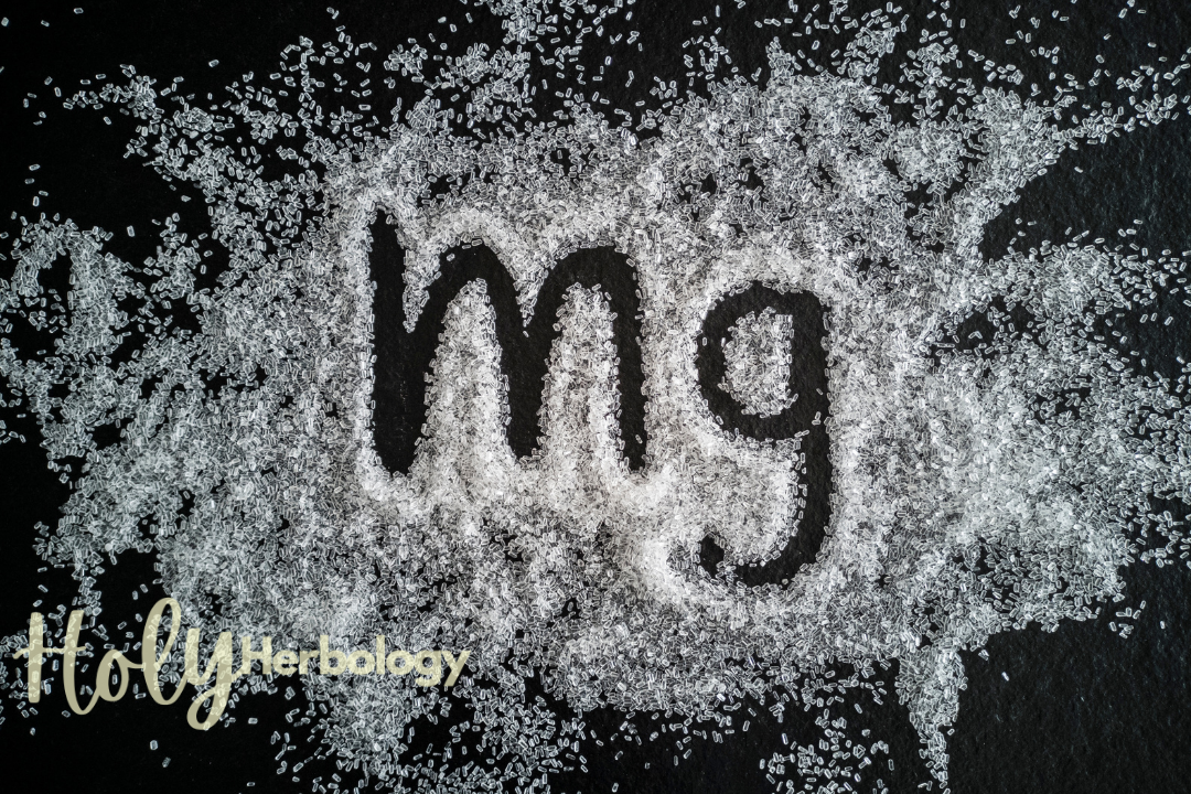 The image features the chemical symbol "Mg" for magnesium, written in a dusting of what appears to be magnesium powder on a dark surface. The phrase "Holy Herbology" is penned in cursive in the bottom left-hand corner, hinting at the importance of this mineral deficiency and its role as a depression-fighting food.