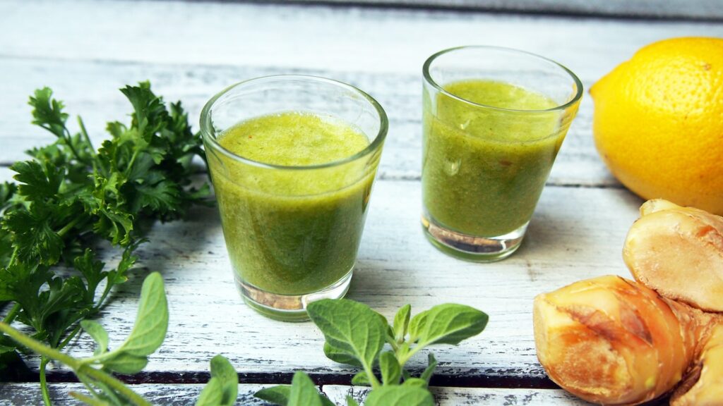 Two small glasses filled with green juice sit on a rustic white wooden surface. Surrounding the glasses are fresh parsley, sprigs of mint, a bright yellow lemon, and chunks of fresh ginger. The scene suggests a healthy, natural drink made from fresh ingredients. Best for daily immunity boost. 