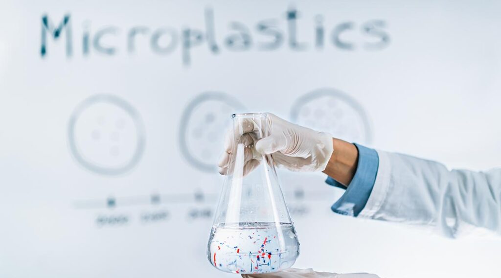 A gloved hand holds a flask filled with water speckled with colorful particles, likely microplastics. The word "Microplastics" and a blurry diagram loom in the background, highlighting the urgent need for eco-friendly solutions to combat plastic exposure.